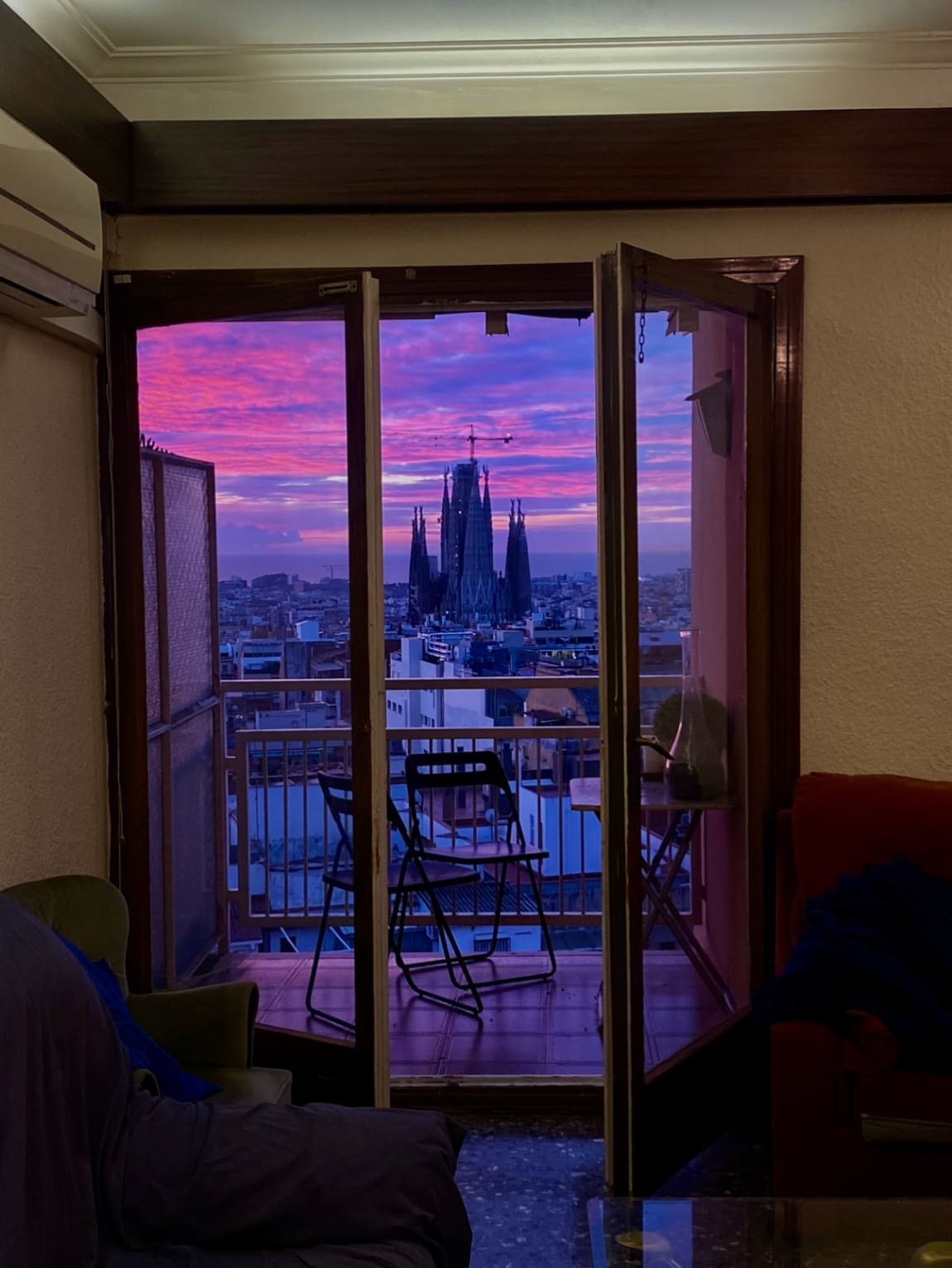 Una porta balconera oberta amb el cel rosa i lila i la silueta de la Sagrada Família al fons