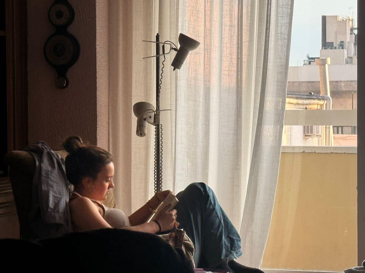 Una noia llegeix un llibre asseguda en una butaca, al costat d’una finestra amb cortines blanques