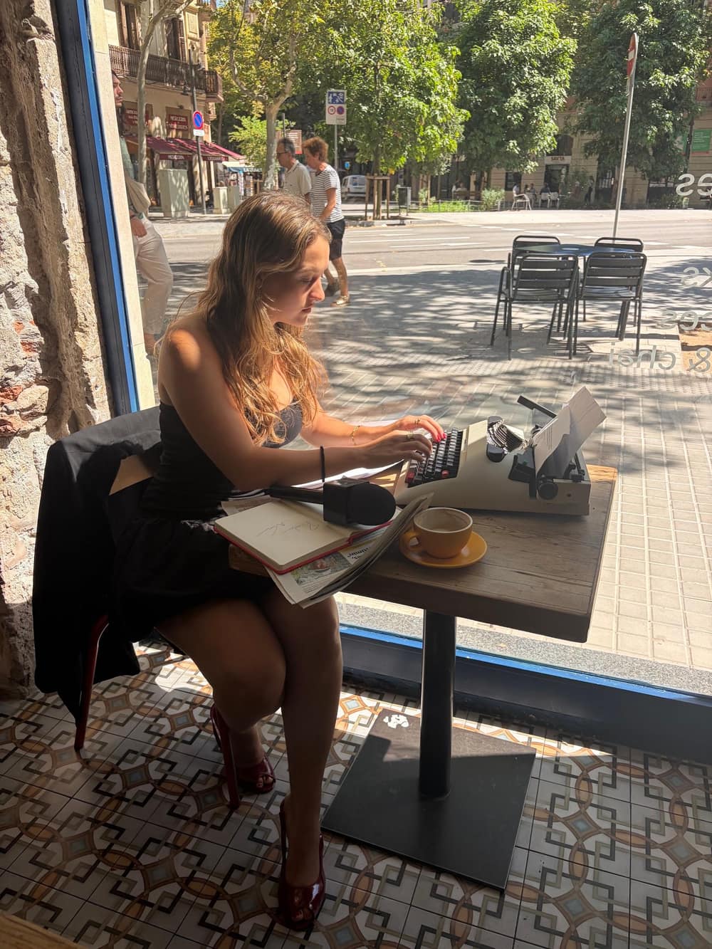 Mireia Riu escriu amb una màquina d’escriure dins d’una cafeteria, al costat de l’aparador, amb un cafè i un micròfon a la taula